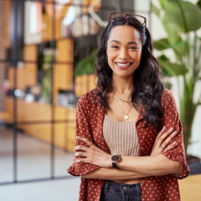 Portrait of smiling young multiethnic woman looking at camera with crossed arms. Successful latin business woman standing in modern office with copy space. Young university hispanic girl with pleasant smile and folded arms.