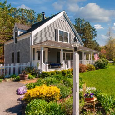An American flag flies from the covered front porch of a  small single family home  with  well maintained grounds and gardens on Cape Cod in Massachusetts.
